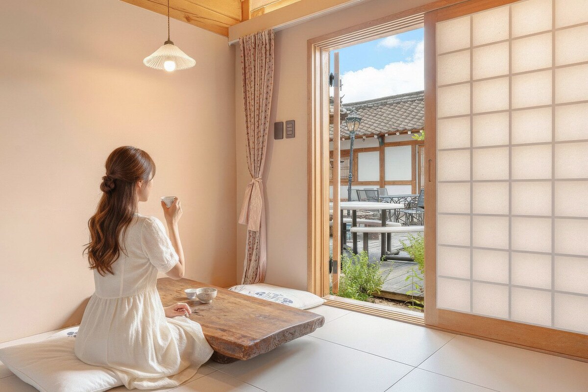 Una habitación con suelo de tatami, una mesa de madera baja y una puerta corredera que da a un espacio exterior con una edificación tradicional japonesa.