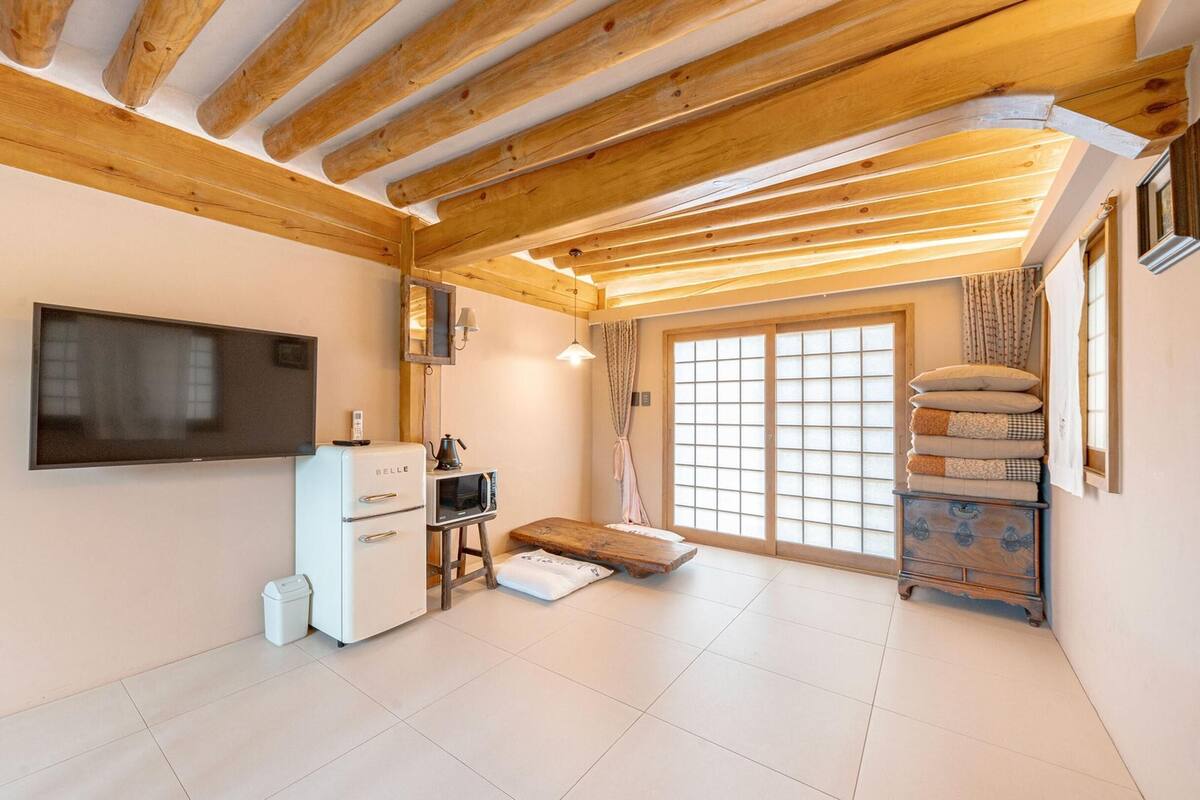 A compact living space with a wooden ceiling, a flat-screen TV, a kitchenette, and a sliding glass door leading to a balcony.