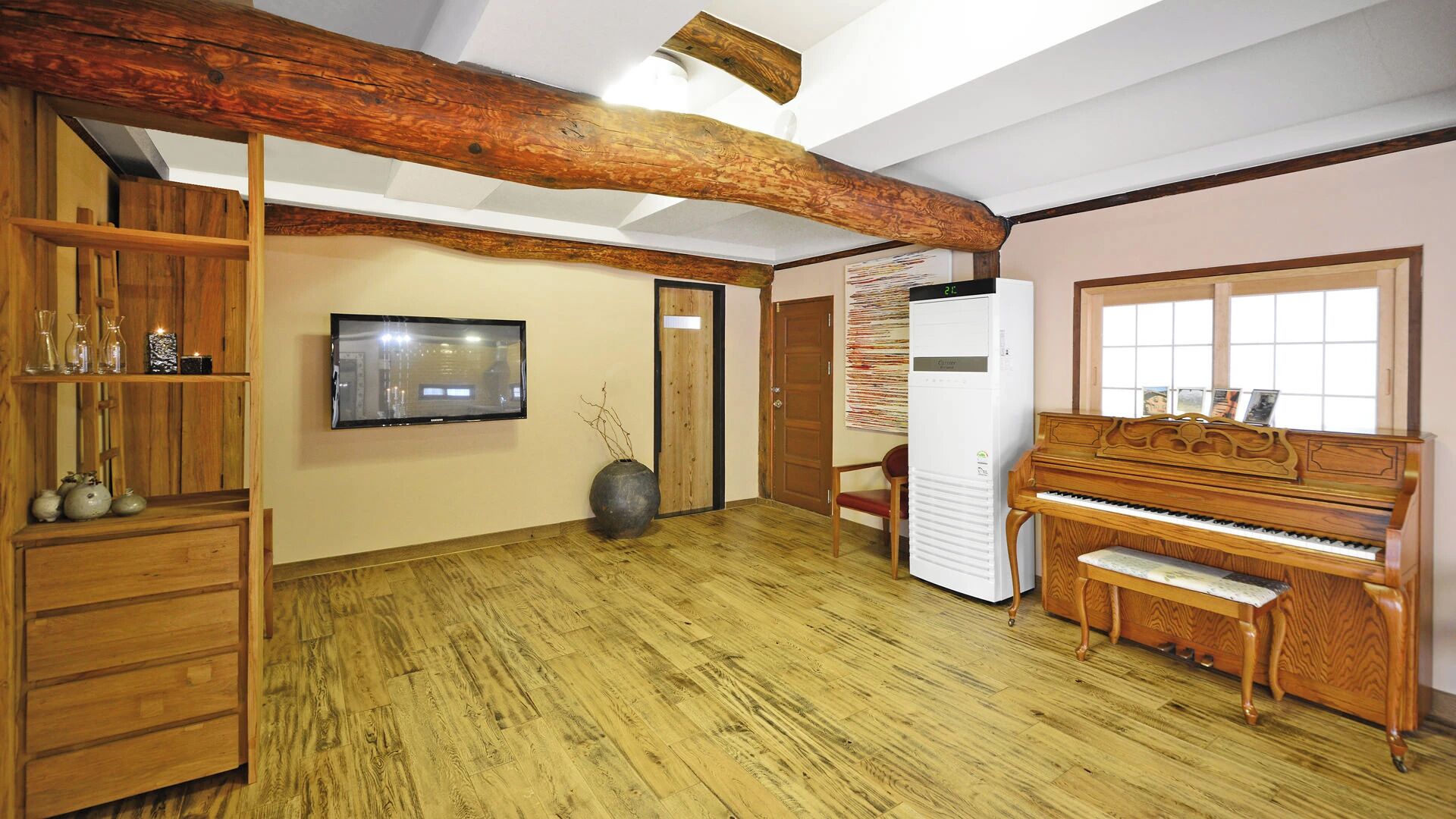 Ein Zimmer mit Holzboden, einem an der Wand befestigten Fernseher, einem Klavier und einem Kühlschrank.