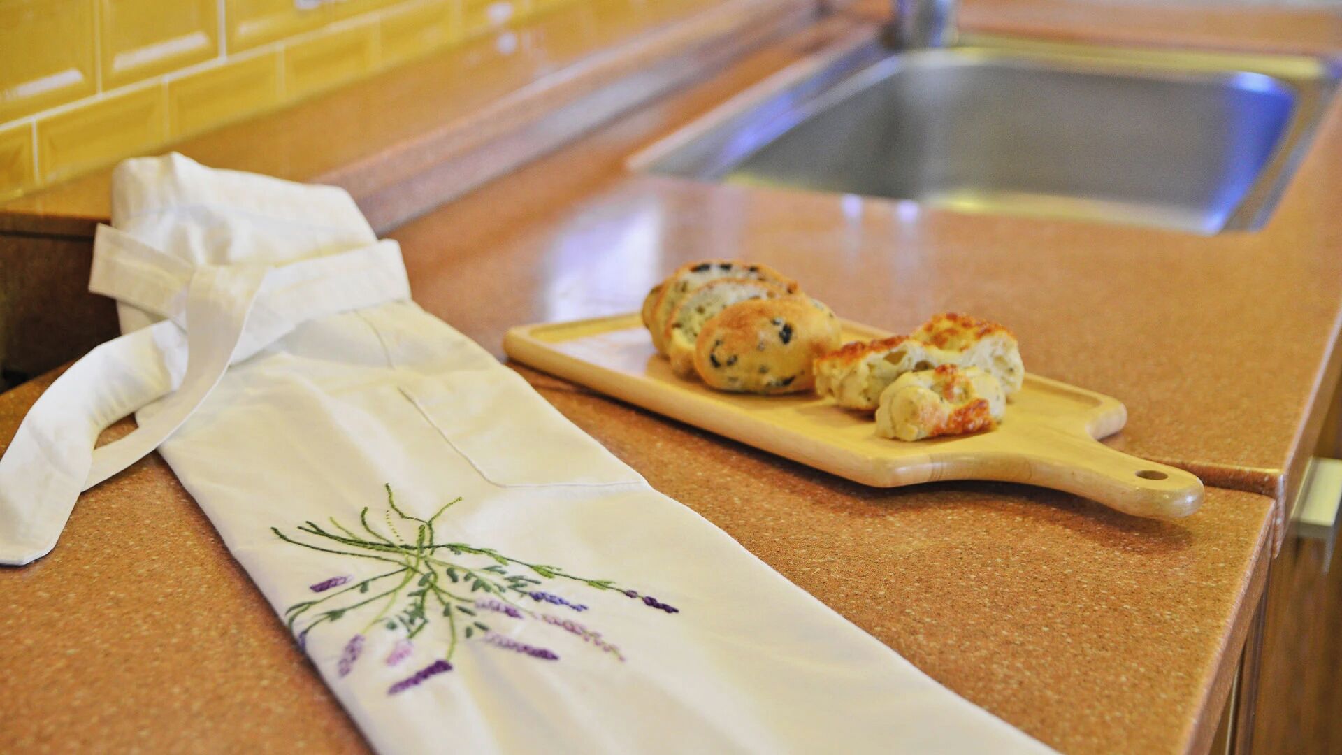 A kitchen counter with a white cloth featuring a floral design and a wooden cutting board with two pastries.
