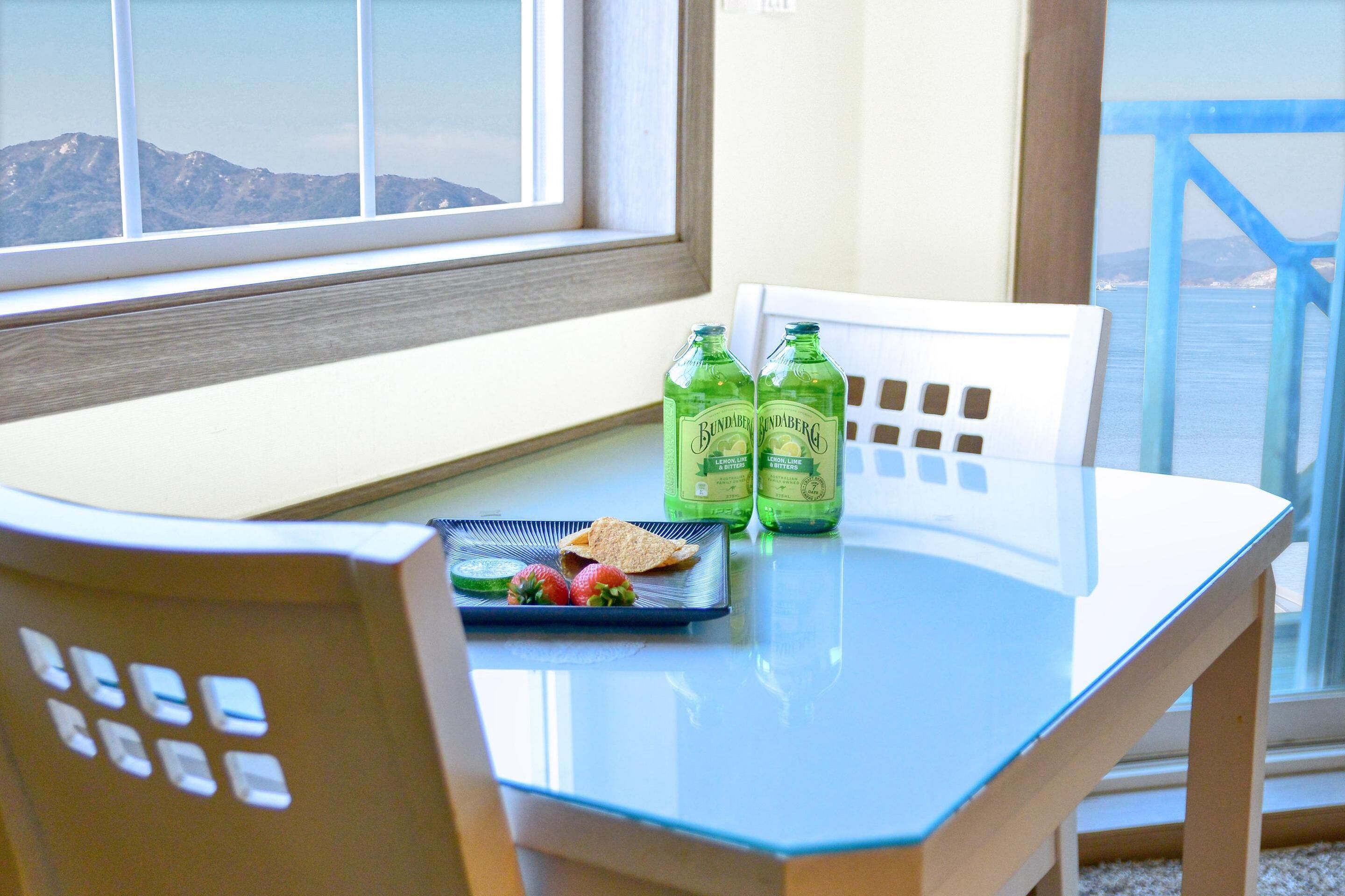 A dining table with two bottles of Bundaberg Ginger Limo, a plate with strawberries and toast, and a view of mountains and the sea through a window.