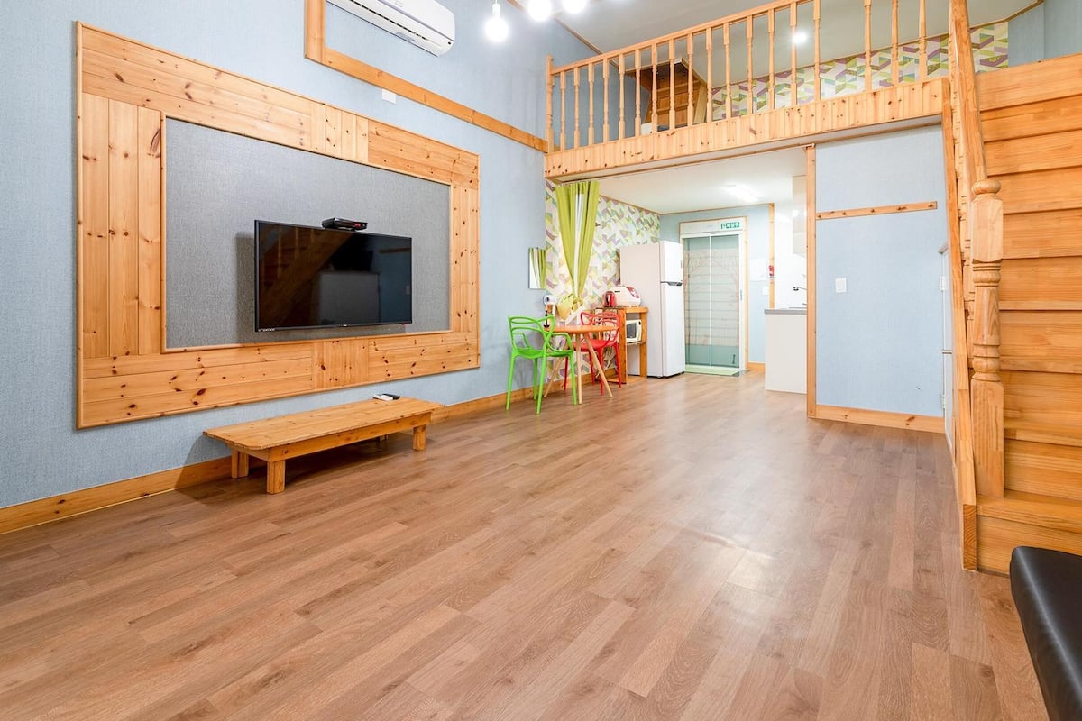 A spacious room with a wooden bench, a TV mounted on the wall, a staircase, and a small table with chairs.