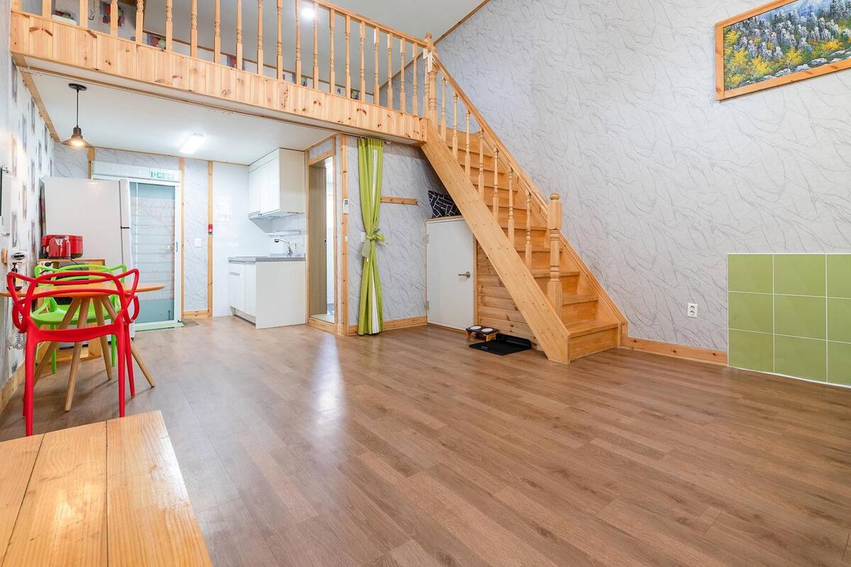 A spacious living area with a wooden staircase, a dining table with chairs, and a kitchen in the background.