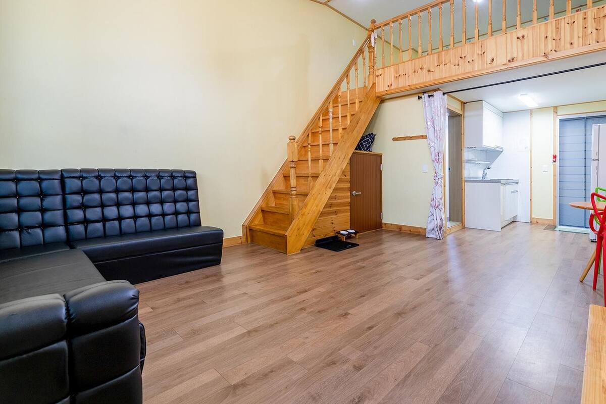 A spacious living area with a wooden staircase, a black leather sofa, and a small kitchen area with a table and chairs.