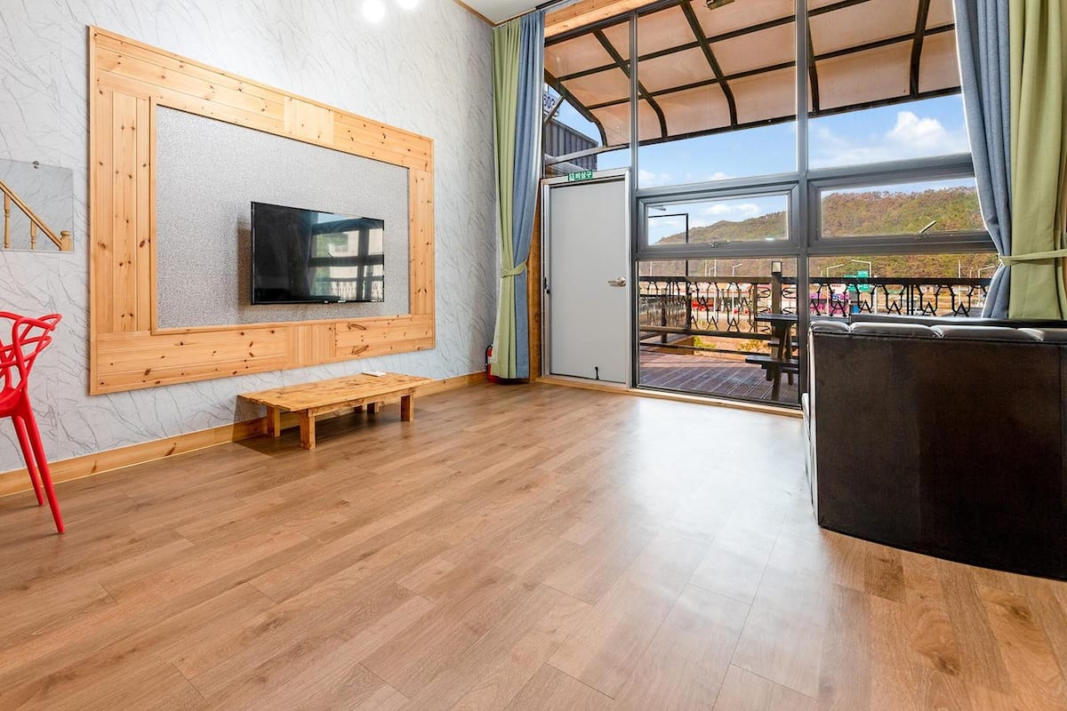 A spacious room with a wooden floor, a TV mounted on the wall, a bench, and a table with a red chair.
