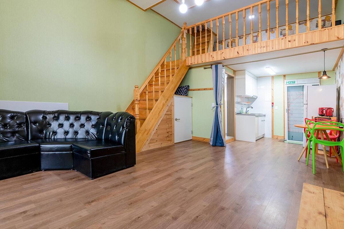 A spacious living area with a black leather sofa, wooden staircase, and a kitchen area with a table and chairs.