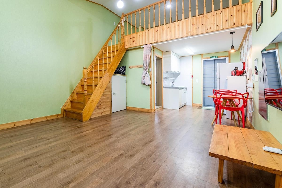 A spacious room with a wooden staircase, a dining area with red chairs, and a kitchen in the background.