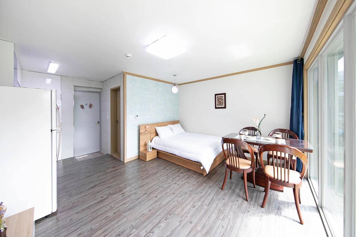 A room with a bed, a dining table with chairs, a refrigerator, and a large window.