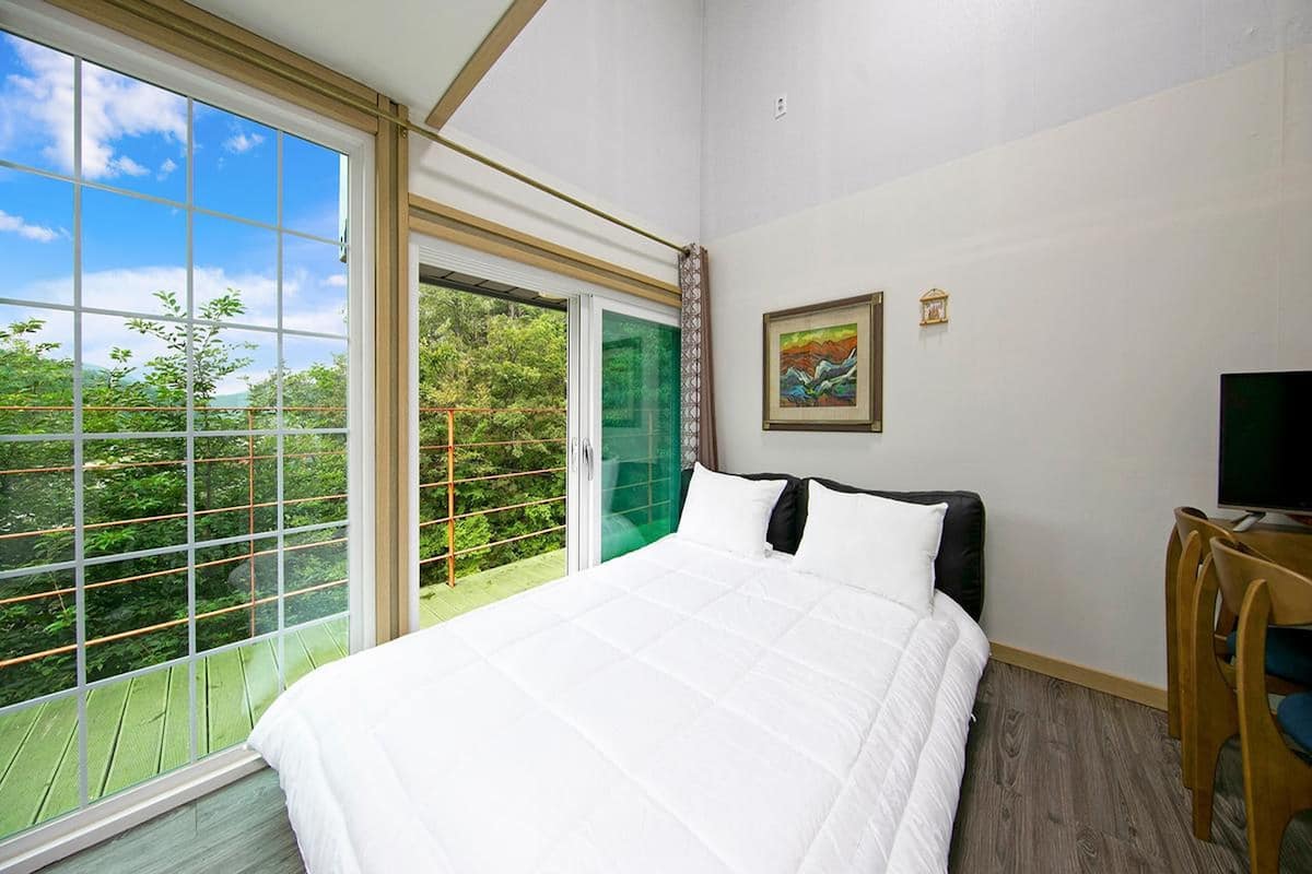 A bedroom with a large window, a bed, a TV, and a painting on the wall.