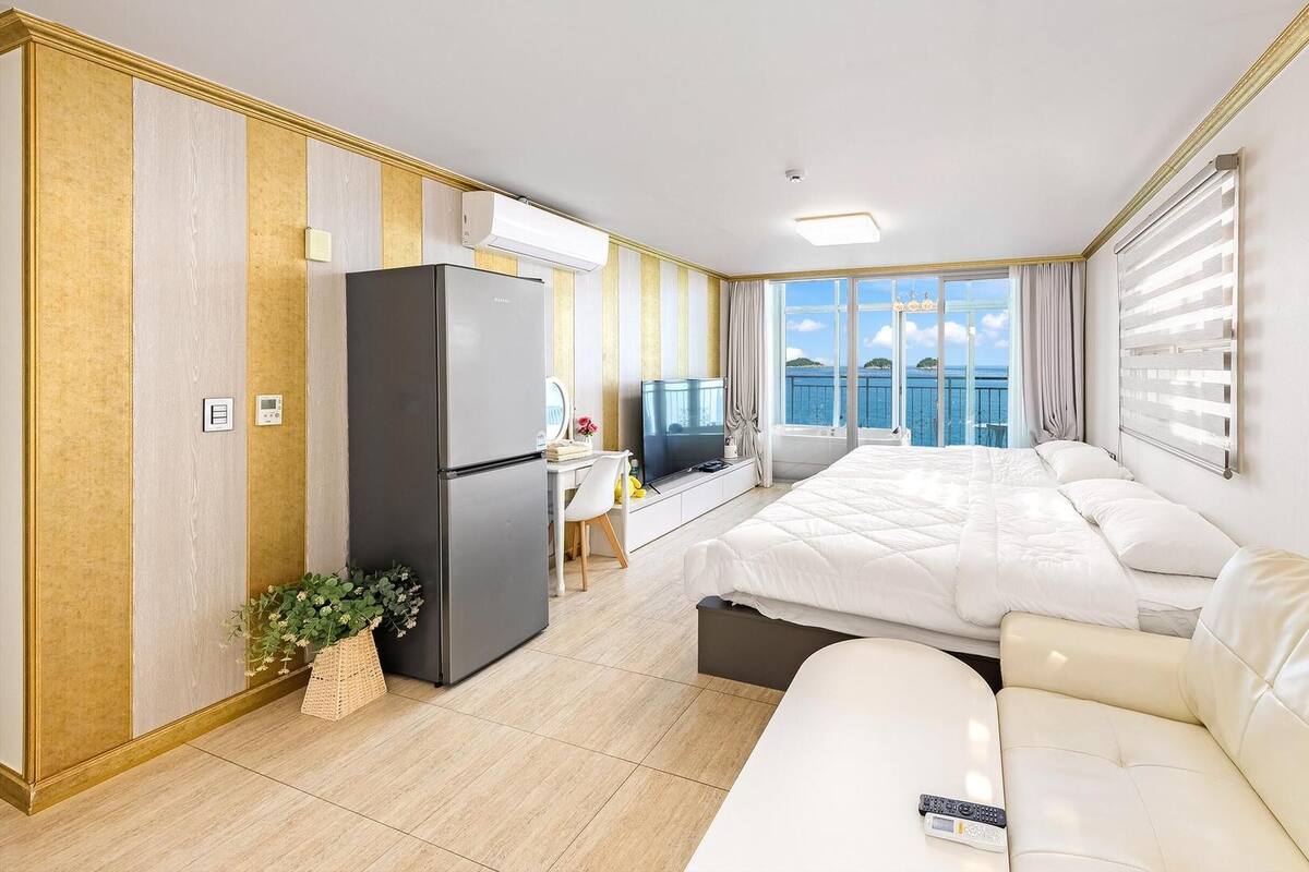Habitación de hotel con cama, sofá, escritorio con televisor, refrigerador y vista al mar.
