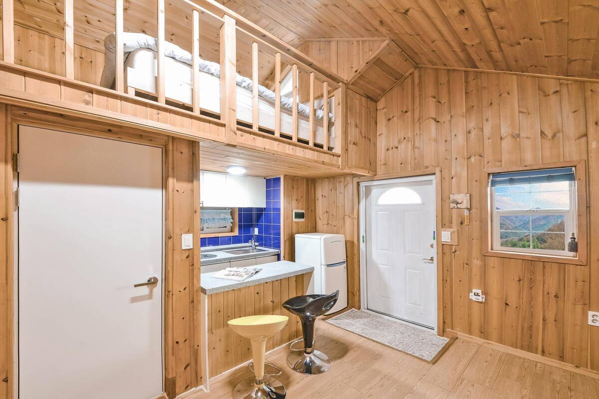 Una cabaña de madera con zona de cocina, comedor con dos taburetes y balcón con cama.