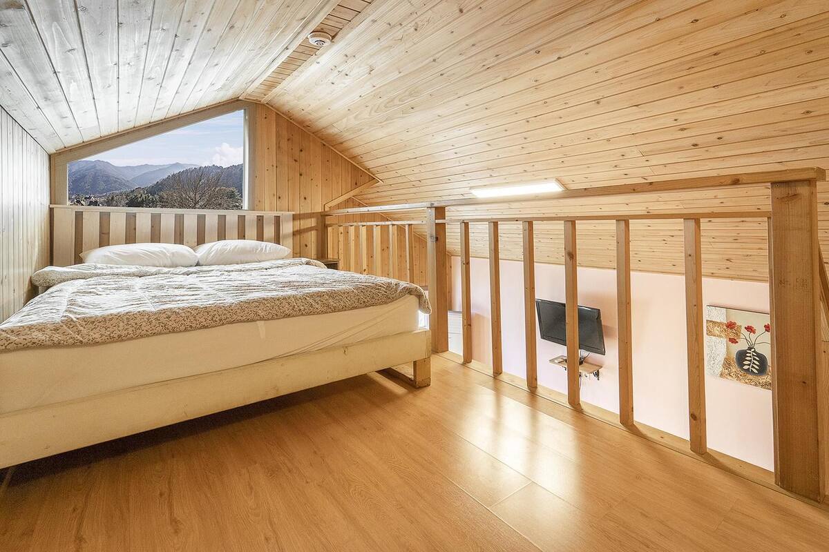 Un dormitorio con techo de madera, cama con ropa de cama blanca, ventana con vista a la montaña y barandilla de madera.