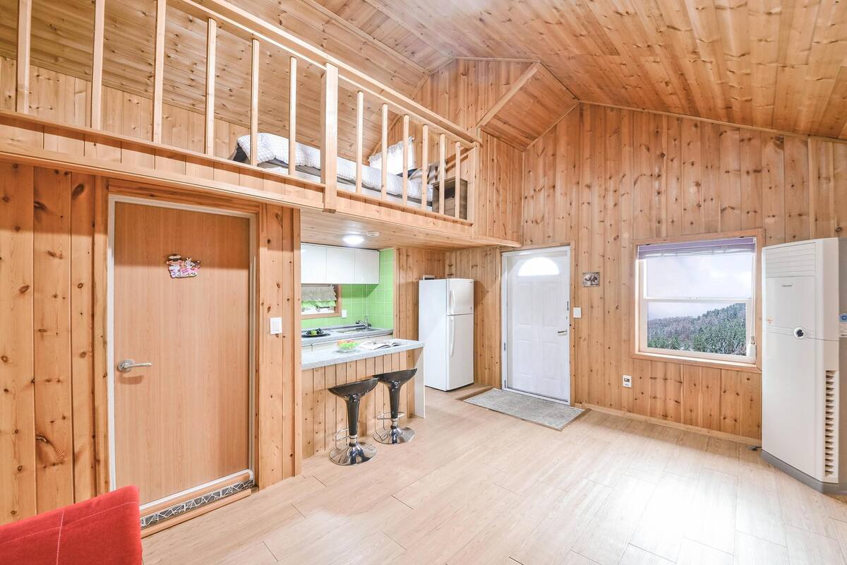 Una cabaña de madera con cocina, una zona de comedor con dos taburetes, un balcón y una ventana con vistas.