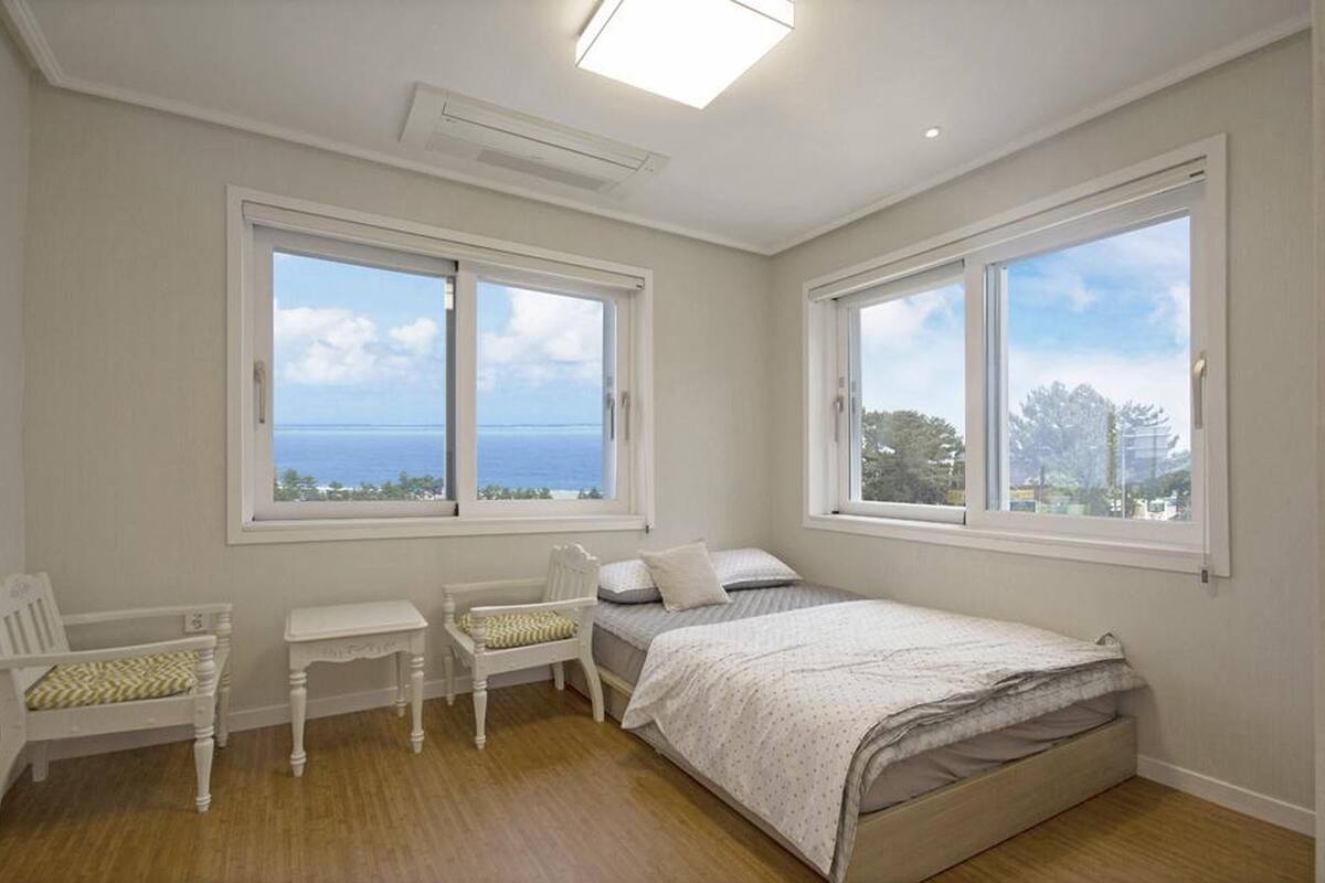 A bedroom with two large windows, a bed, a chair, and a small table.