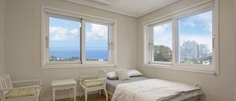 A bedroom with two large windows, a bed, a chair, and a small table.