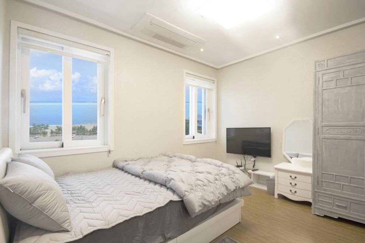 A bedroom with a bed, a TV, a dresser, and a view of the sea.