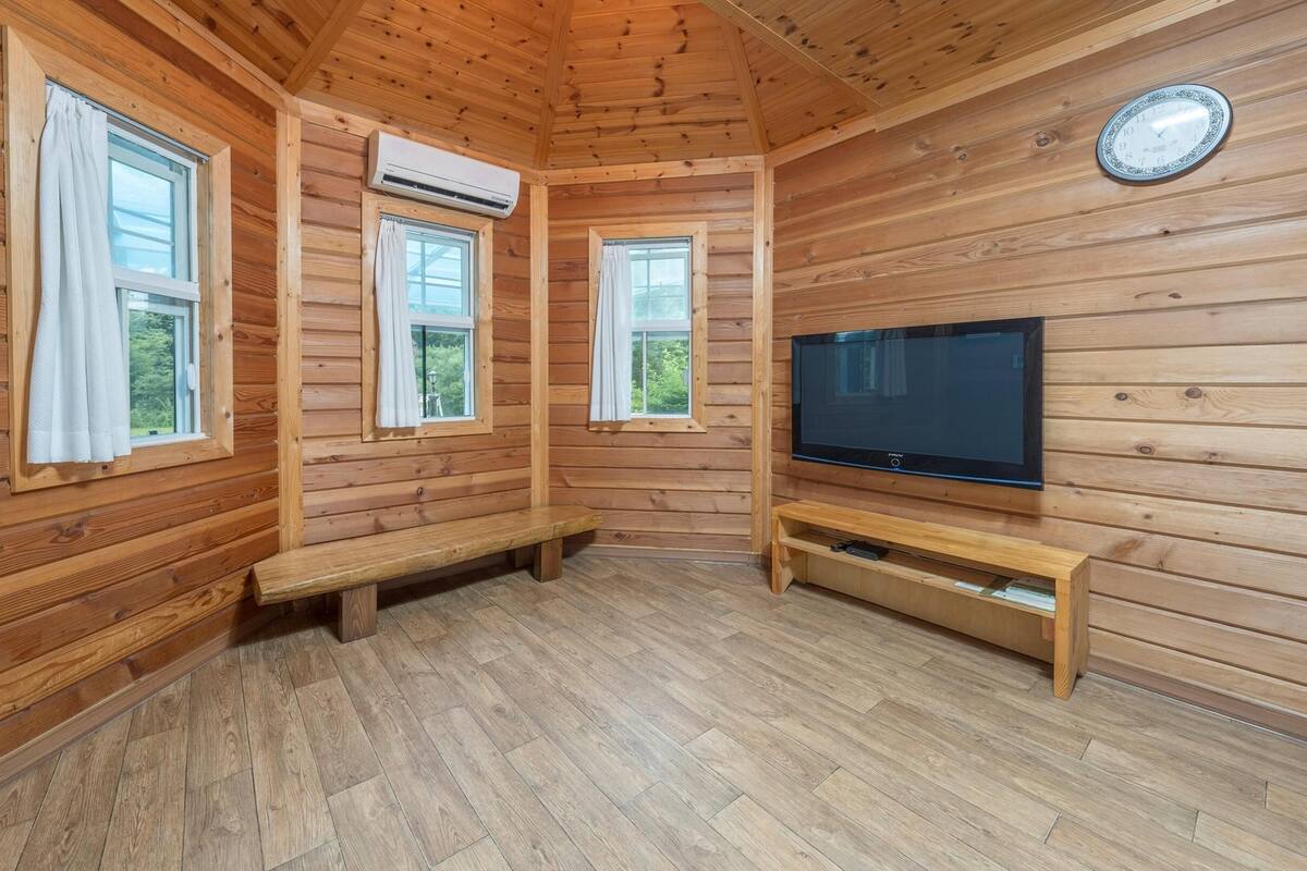 A wooden cabin room with a TV, bench, and three windows.