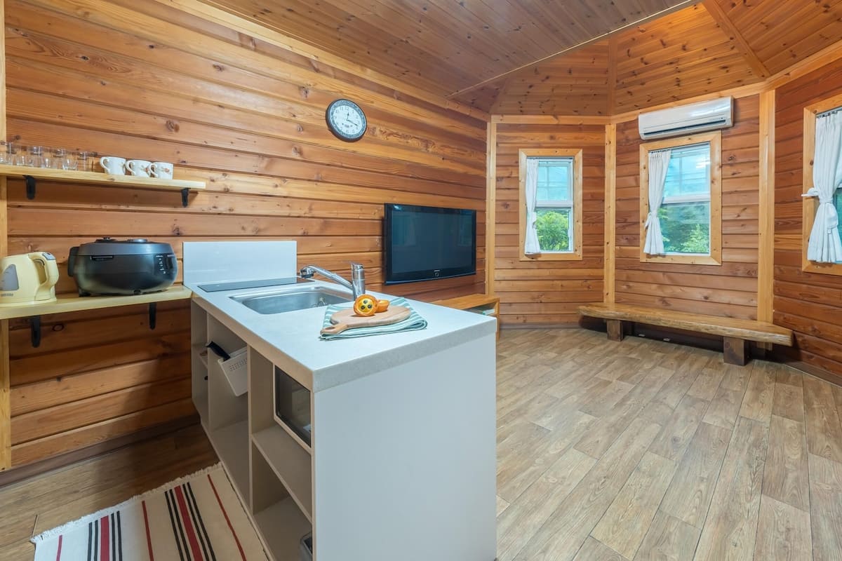 A compact kitchen with wooden walls, a sink, a clock, and two windows.