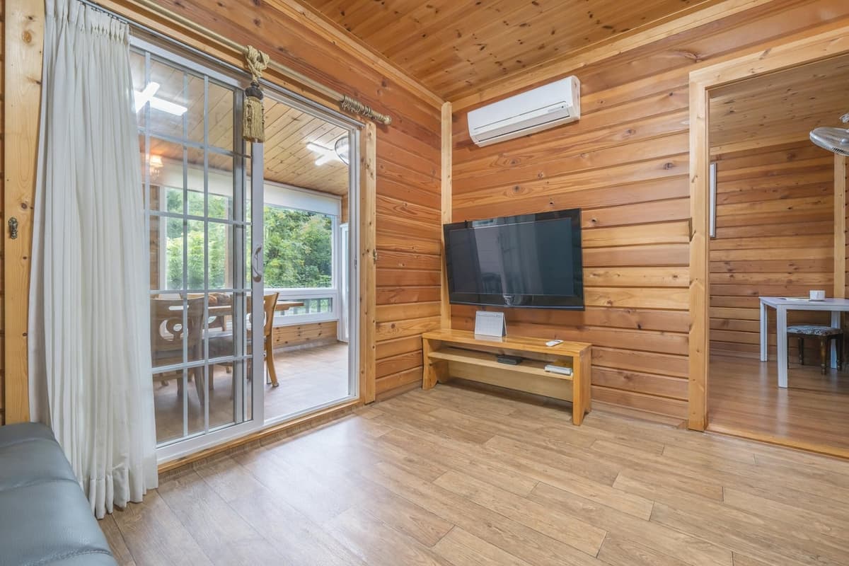 A wooden-paneled room with a sliding glass door leading to an outdoor area, a flat-screen TV mounted on the wall, and a small table with a chair.