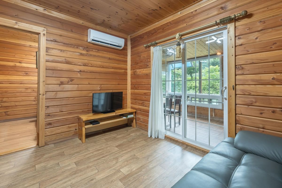A wooden cabin interior with a television on a wooden stand, a sofa, and a sliding glass door leading to an outdoor deck.
