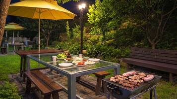 An outdoor dining area with a table set for a meal, surrounded by greenery and a barbecue grill.
