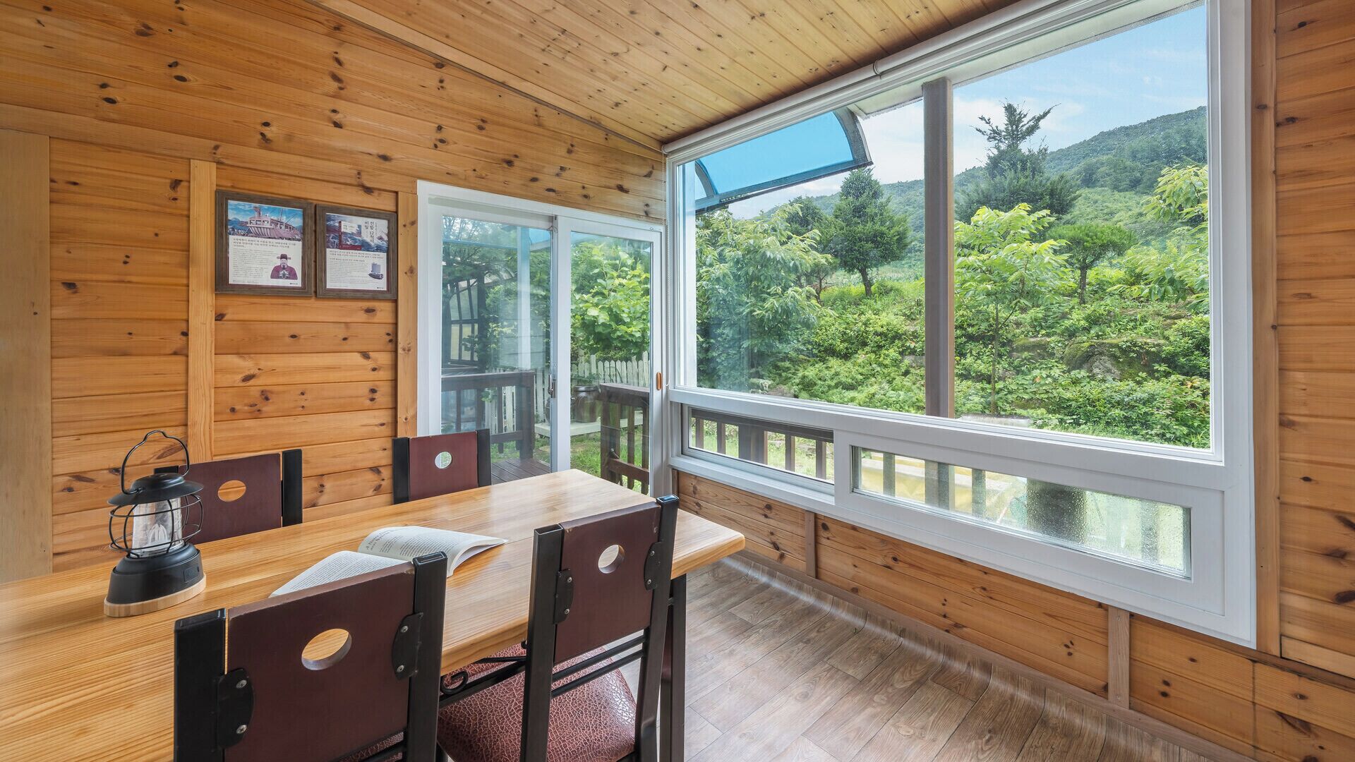 A wooden cabin with a dining area, large windows, and a view of a forested landscape.