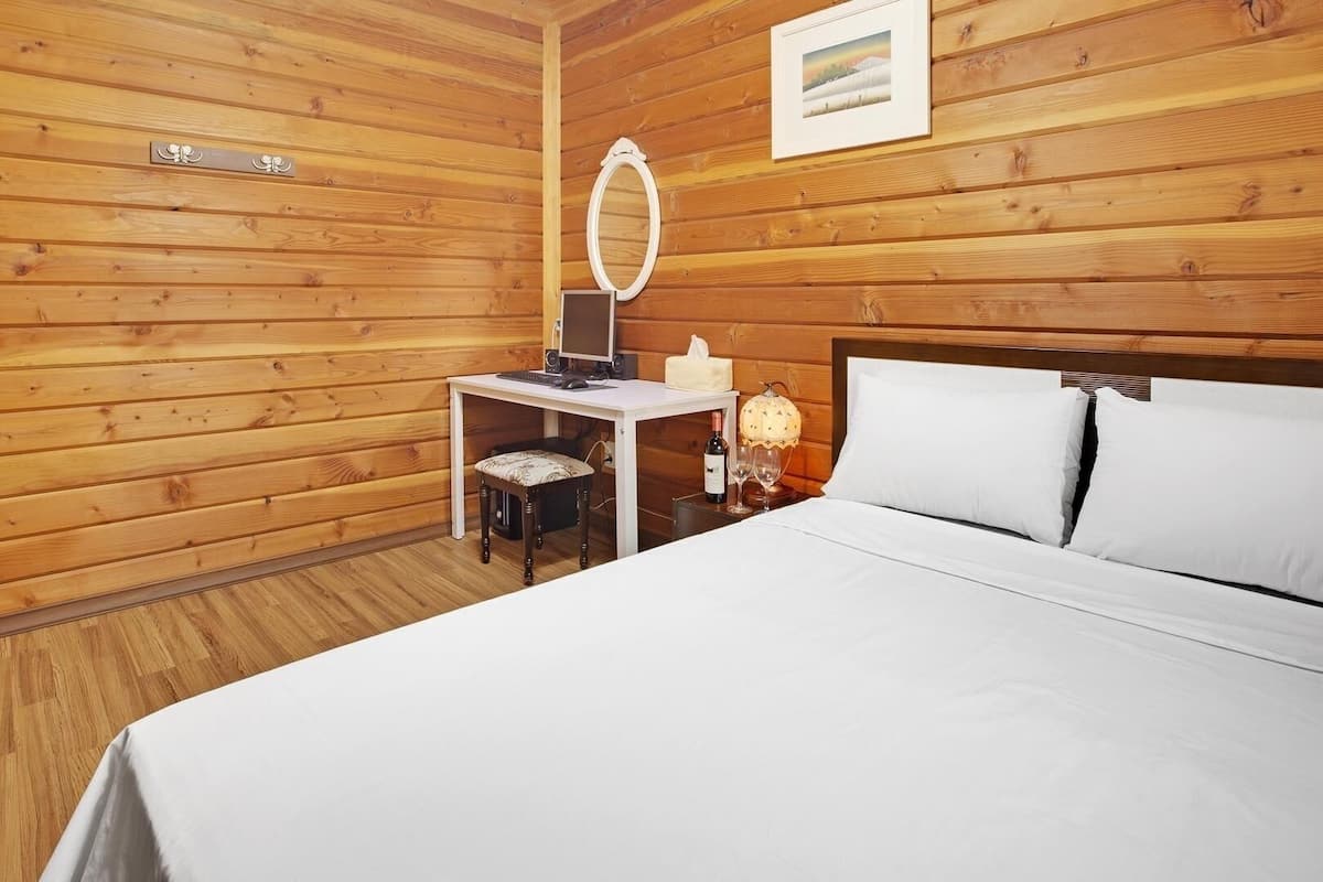 A wooden-paneled room with a bed, a desk with a computer, a chair, a lamp, and a mirror.