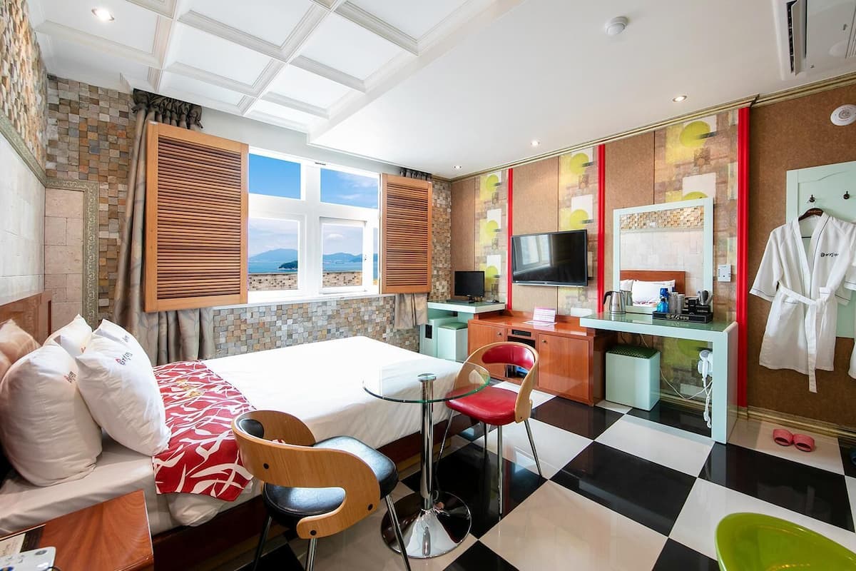 A hotel room with a bed, a desk with a TV, a chair, a red chair, a mirror, and two robes on a rack.