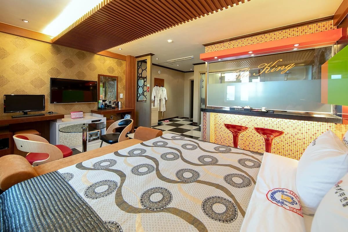 A hotel room with a bed, a desk, a TV, a mirror, and a bathroom with a sink and red bar stools.