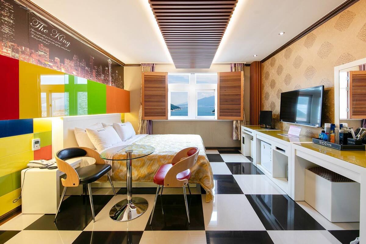 A hotel room with a bed, a desk with a TV, a window with a view, and a colorful wall.