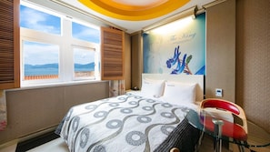 A hotel room with a bed, a window with a view of the sea, a wall with a painting, and a glass table with a red chair. - Yeosu King Pension Tel (Yeosu)