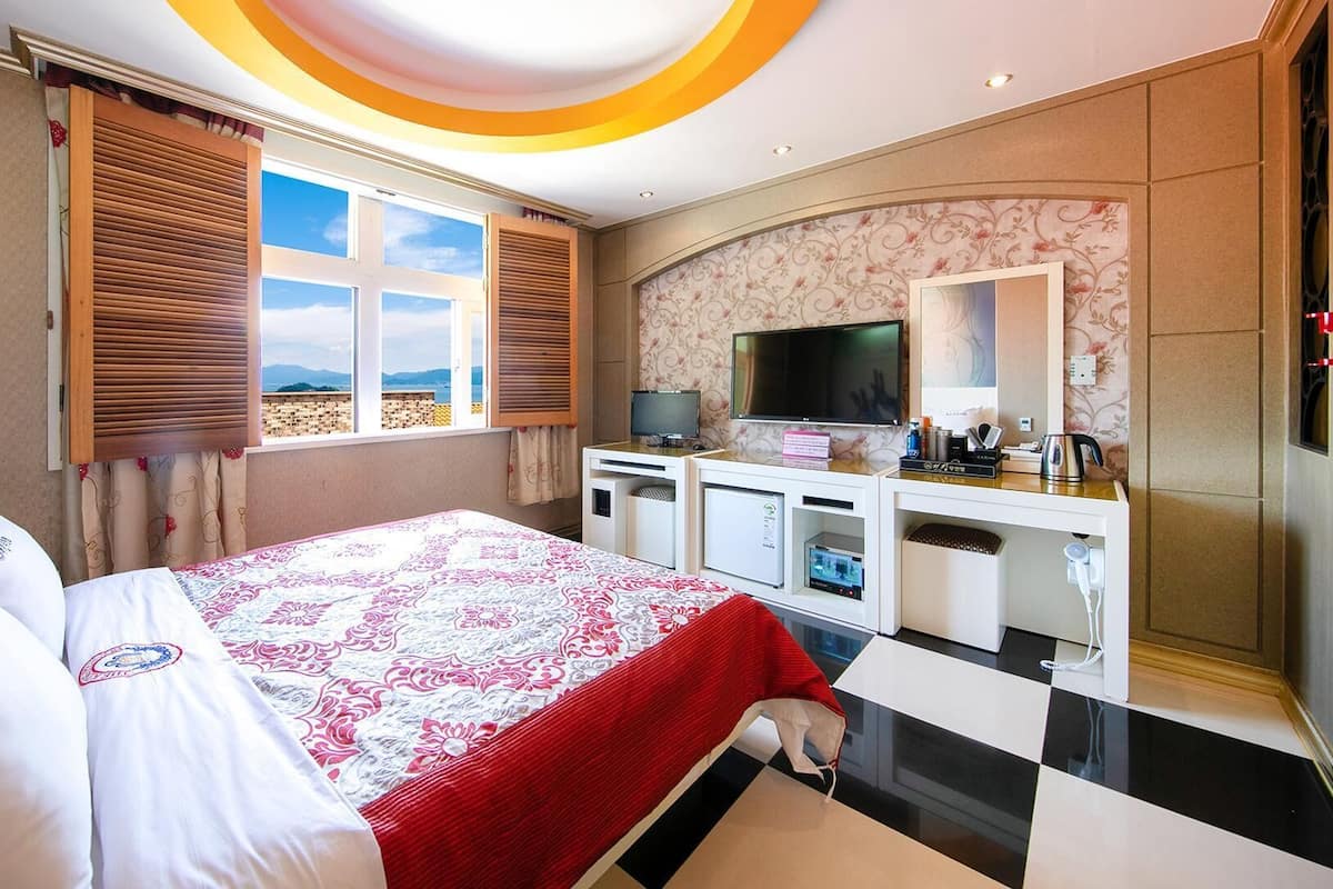 A hotel room with a bed, a desk, a TV, and a view of the beach.