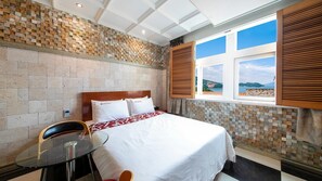 A hotel room with a bed, a glass table, and a view of the sea through a window with wooden shutters. - Yeosu King Pension Tel (Yeosu)
