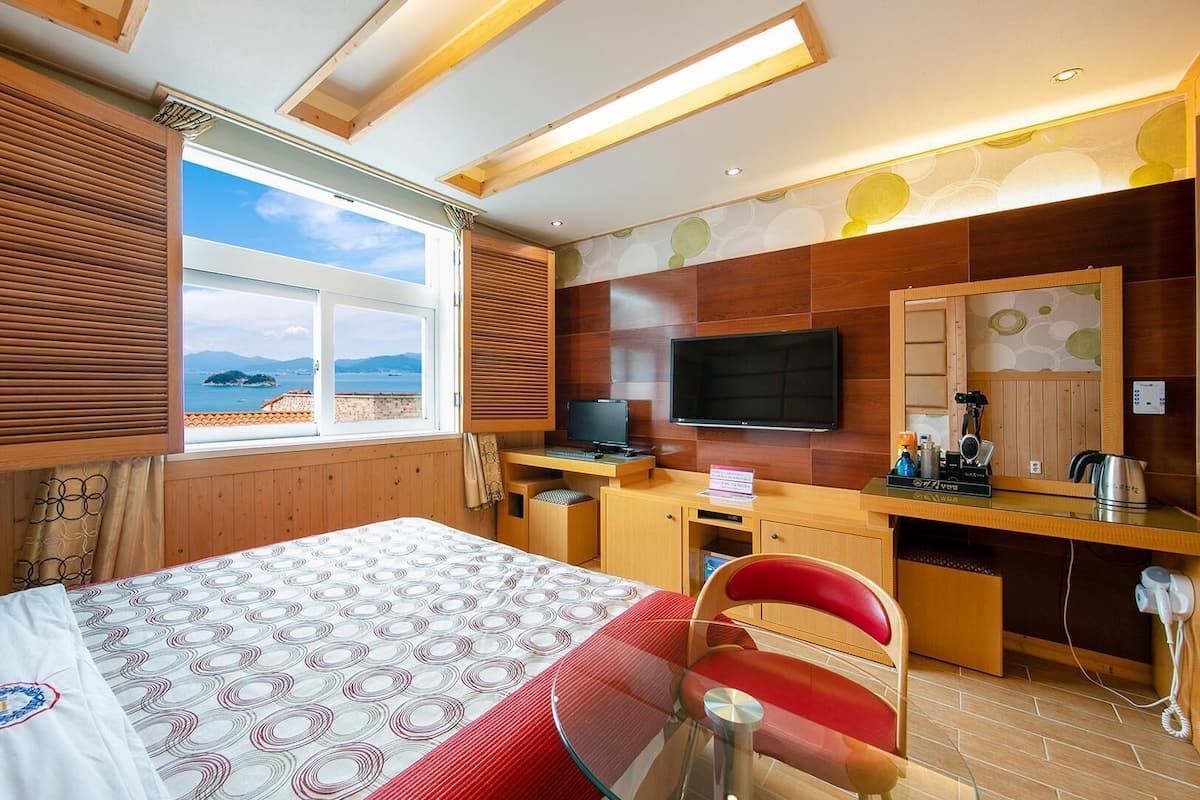 A hotel room with a bed, a desk with a computer, a TV, and a view of the sea.