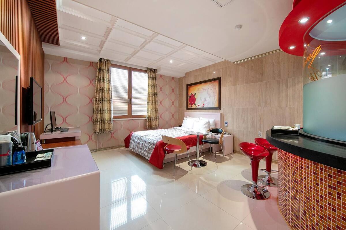 A modern hotel room with a bed, a desk with a computer, a TV, and a bathroom with a red sink.