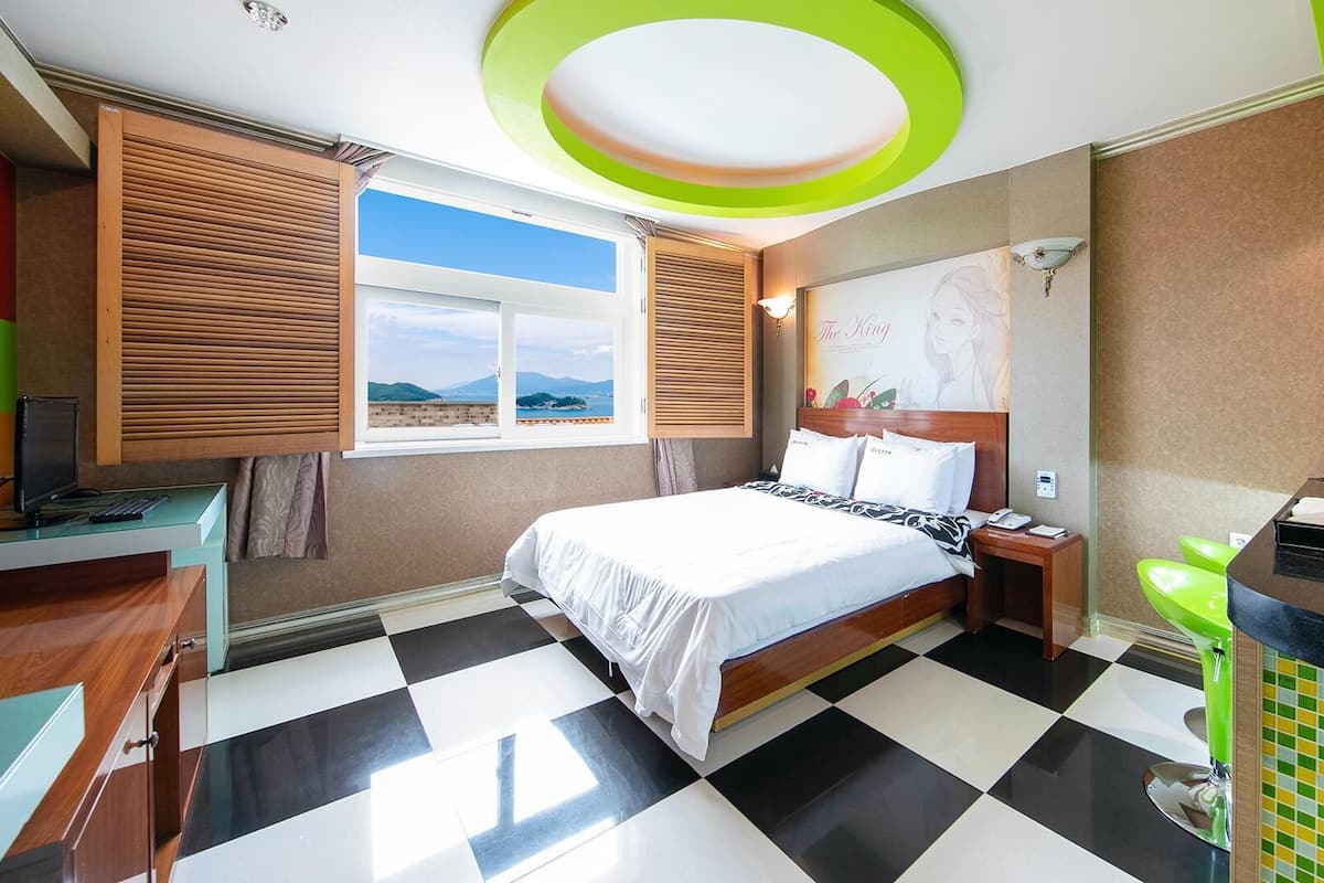 A hotel room with a checkered floor, a bed with white linens, a desk with a computer, and a window with a view of the mountains.