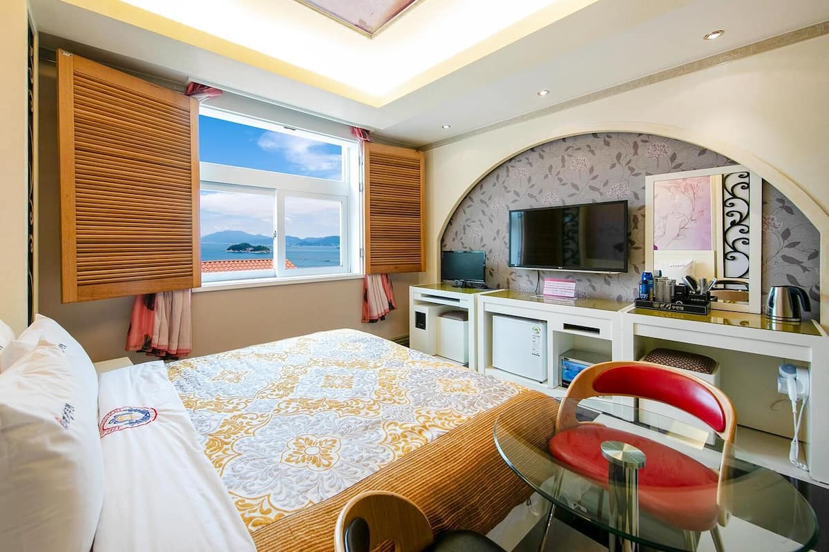 A hotel room with a bed, a desk with a computer, a TV, and a view of the sea.