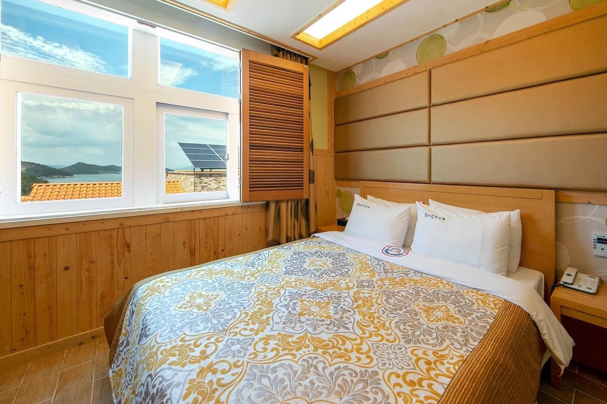A bedroom with a large bed, wooden headboard, a nightstand with a phone, and a window with blinds.