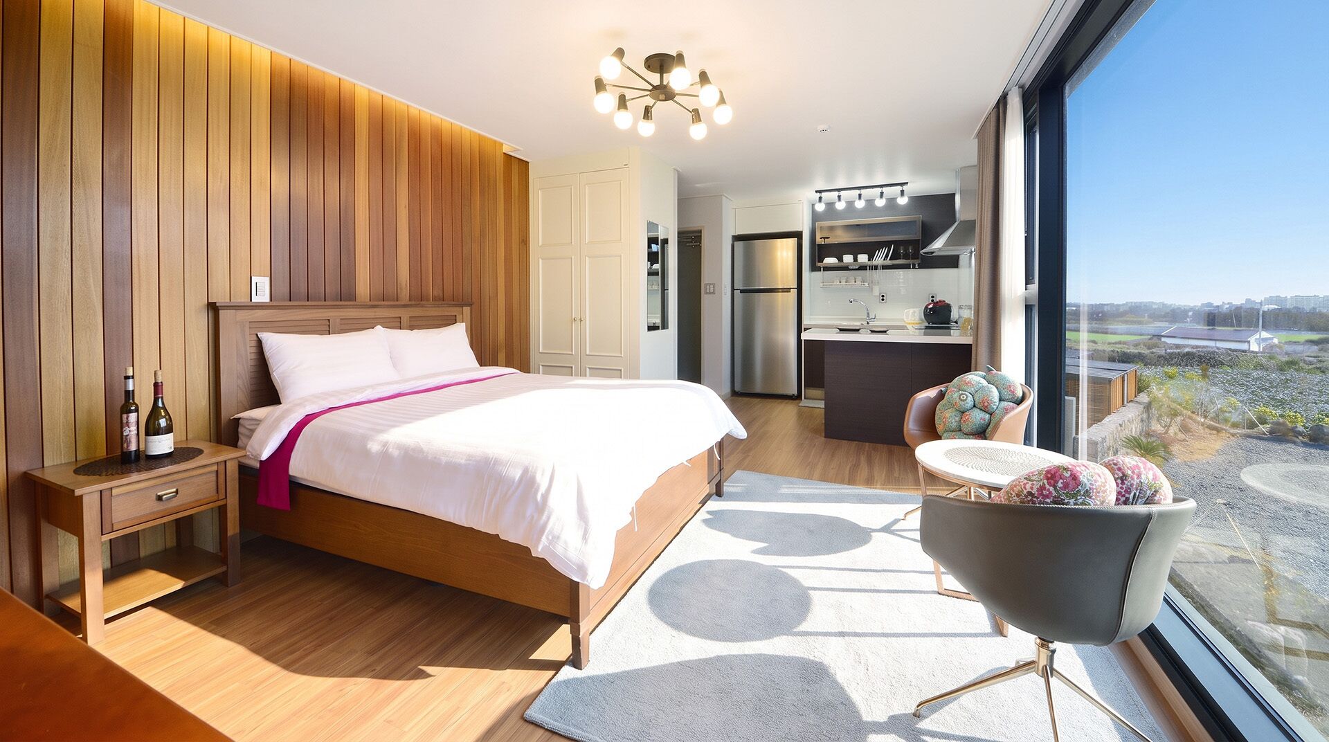 Un dormitorio moderno con una cama grande, mesitas de noche, un pequeño tocador, vistas a la cocina y un balcón con una silla.