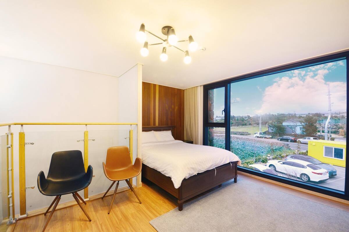 Ein Schlafzimmer mit großem Fenster, einem Bett, zwei Stühlen und einer Deckenleuchte.