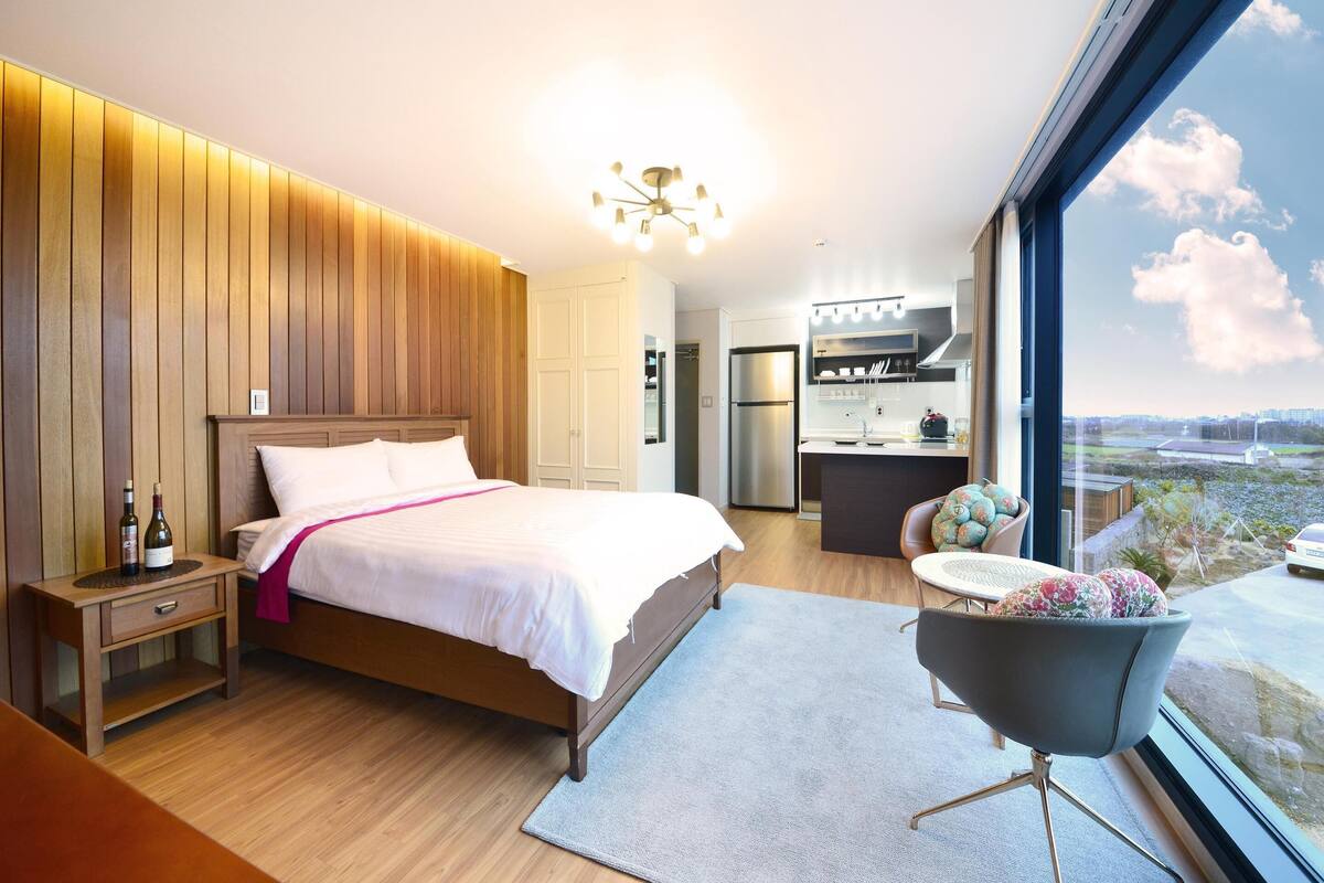 Ein modernes Schlafzimmer mit einer Holzpaneelwand, einem großen Bett, einem Nachttisch, einem Sessel und Blick auf die Stadt durch ein großes Fenster.