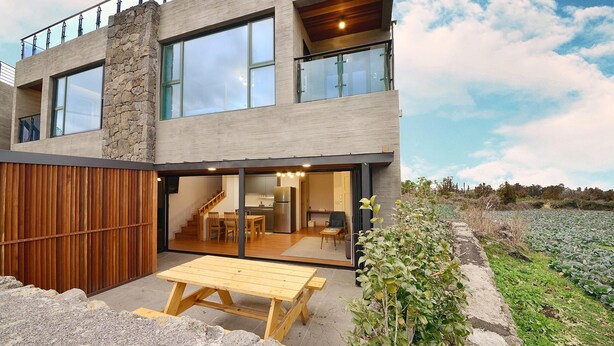 Une maison moderne à deux étages, dotée d’une façade en pierre, de grandes fenêtres et d’une terrasse en bois avec une table de pique-nique.