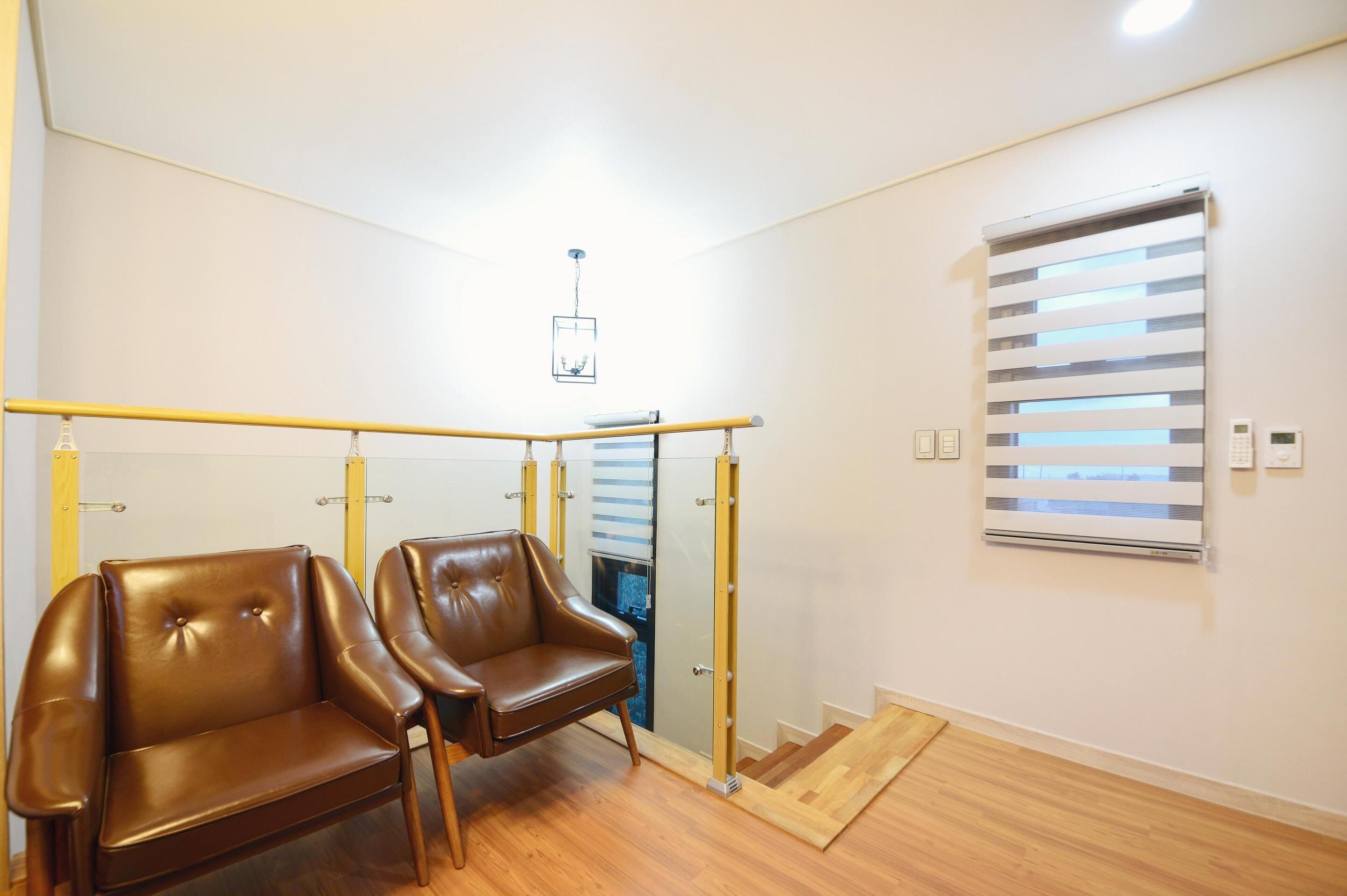 Een kamer met twee bruine leren fauteuils, een houten vloer, een plafondlamp, een raam met jaloezieën en een wandairconditioning.