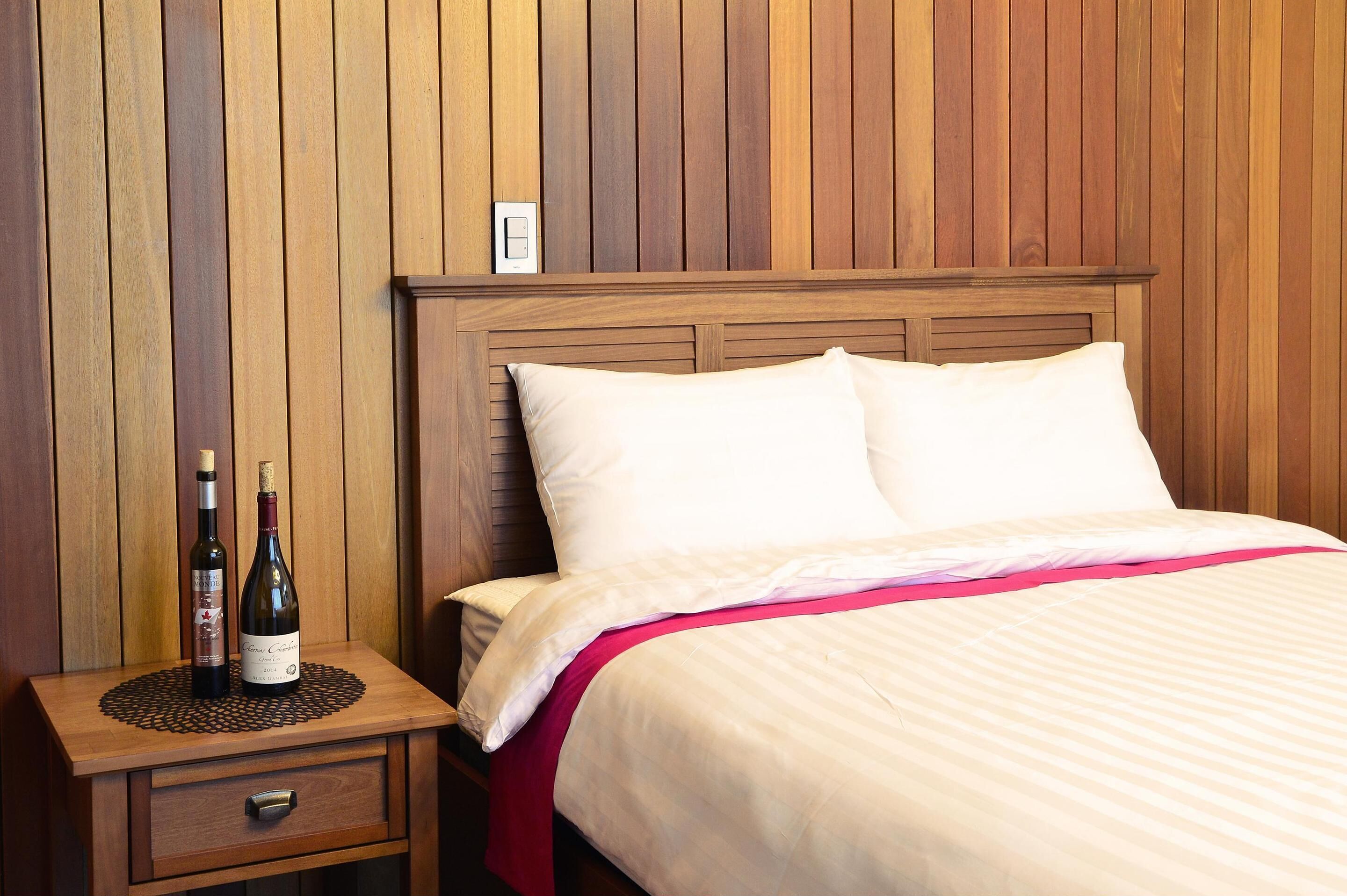 A neatly made bed with a wooden headboard, a bedside table with two bottles of wine, and a small bedside lamp.
