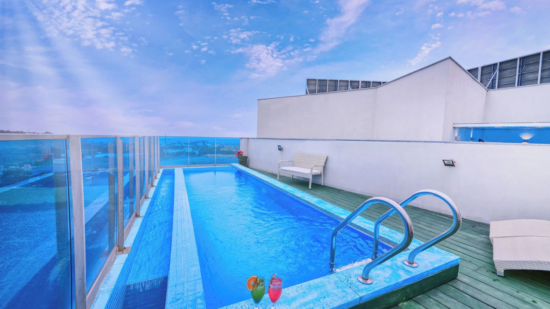 A rooftop infinity pool with clear glass railings, a white bench, and a wooden deck.