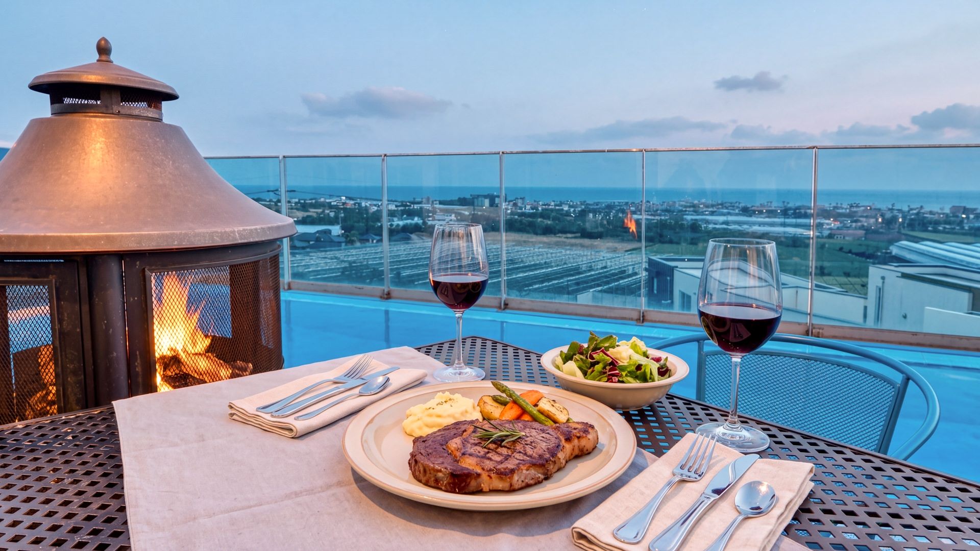 Une table à manger avec un steak, une salade et deux verres de vin rouge, donnant sur un paysage urbain.