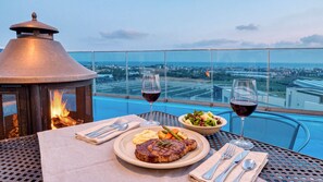 Une table à manger avec un steak, une salade et deux verres de vin rouge, donnant sur un paysage urbain.