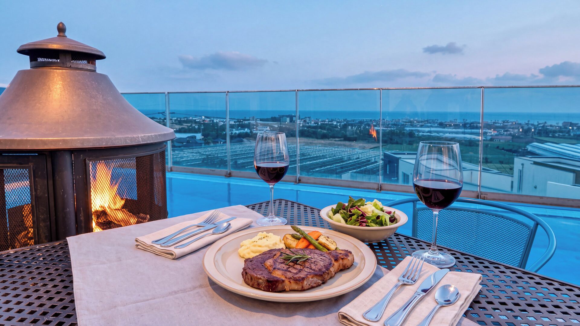 Une table à manger avec un steak, une salade et deux verres de vin rouge, donnant sur un paysage urbain.