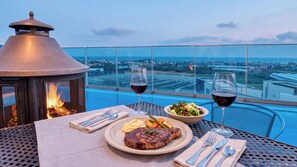 Una mesa de comedor con un bistec, ensalada y dos copas de vino tinto, con vistas a un paisaje urbano.
