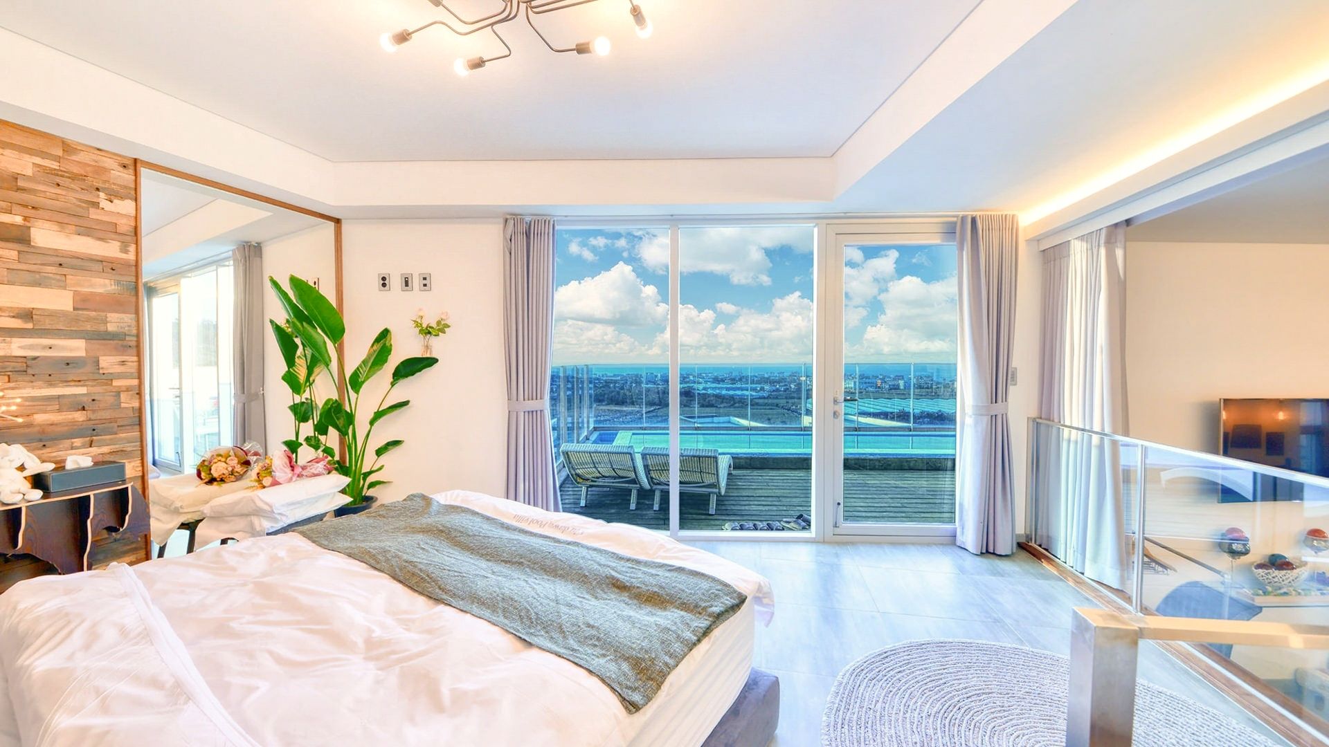 Ein modernes Schlafzimmer mit einem großen Bett, Blick auf ein Schwimmbad und eine Stadtlandschaft durch raumhohe Fenster sowie ein Balkon mit Glasgeländer.